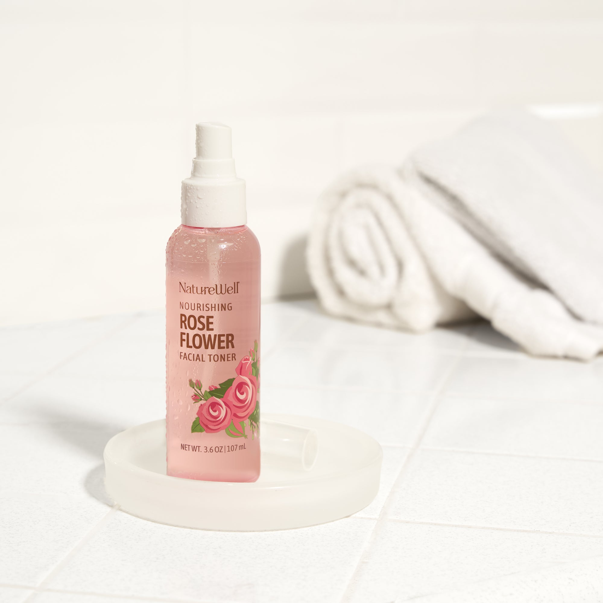 3.6 oz bottle of nourishing rose flower facial toner spray on white tiled bathroom counter aside a white towel