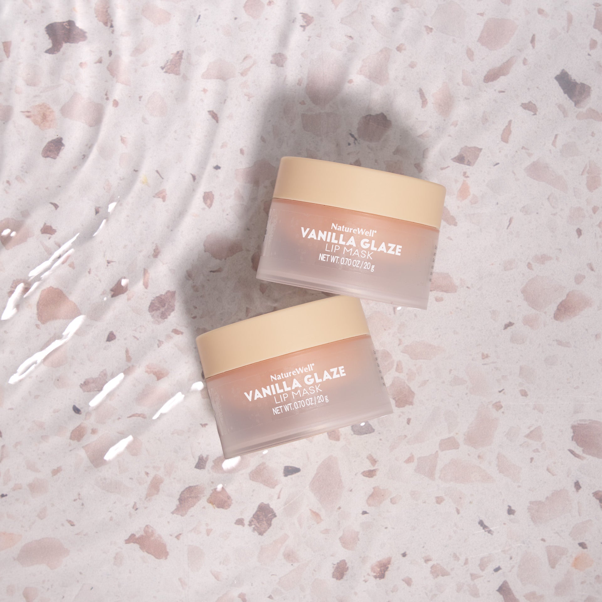 2 pale pink jars of vanilla glaze lip mask atop a neutral stone patterned background