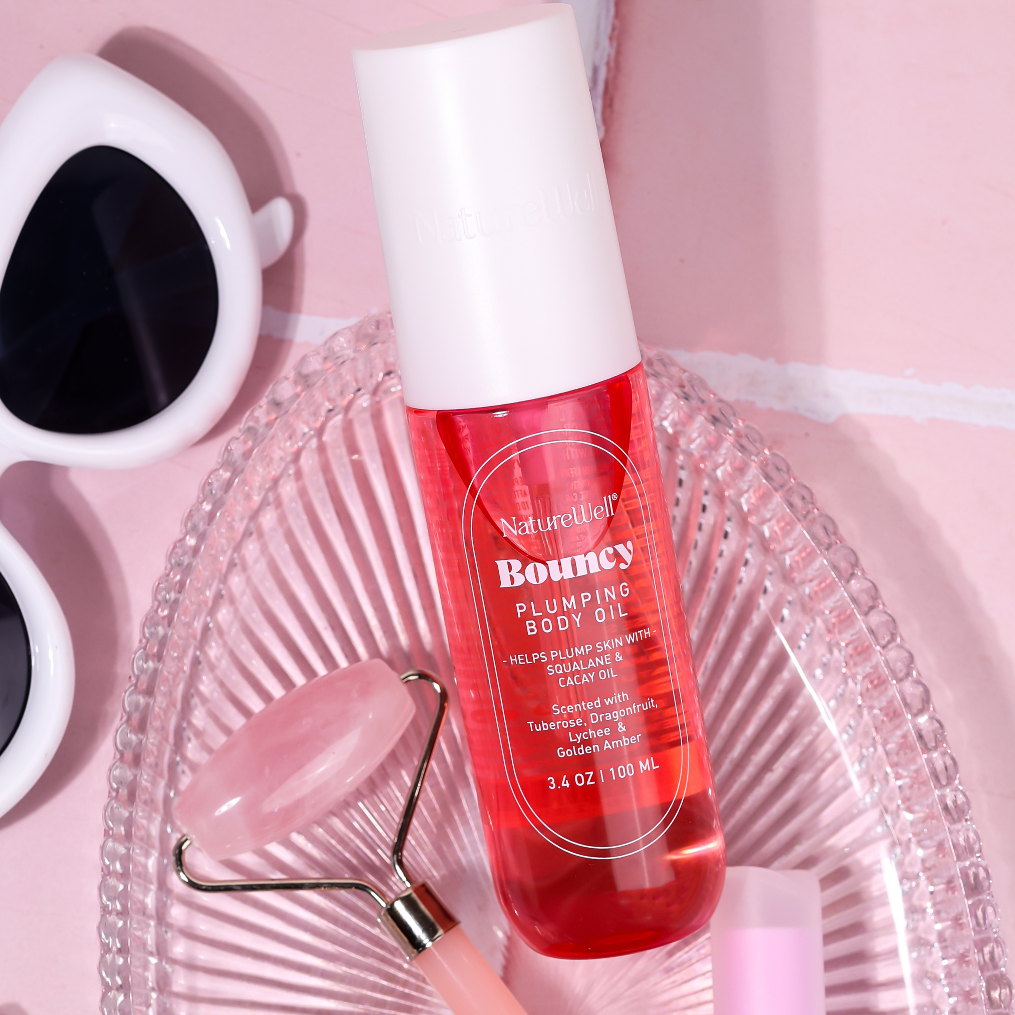 Bottle of NatureWell Bouncy Plumping Body Oil on a pink tile countertop with other beauty products.