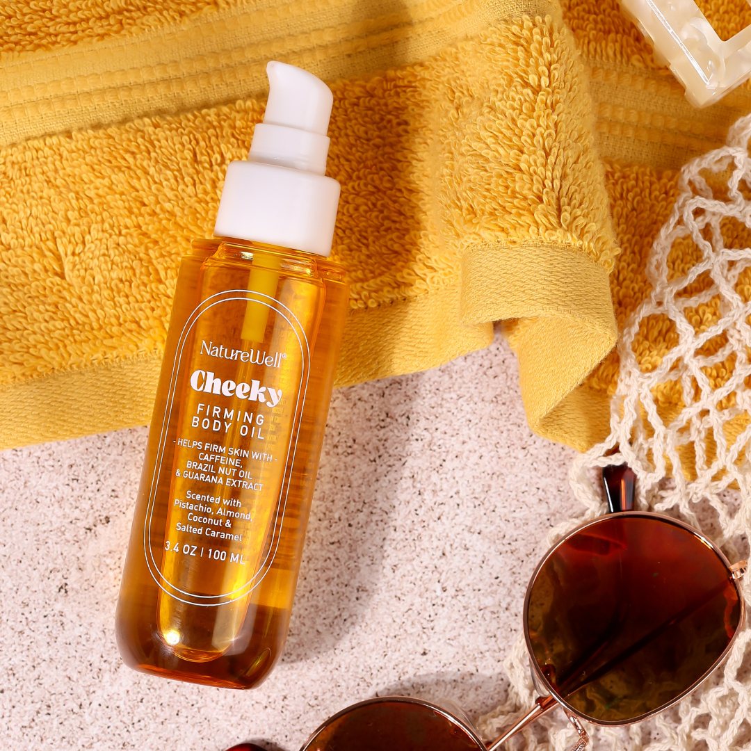 Bottle of NatureWell Cheeky Firming Body Oil on a sandy beach surrounded by a beach towel and sunglasses