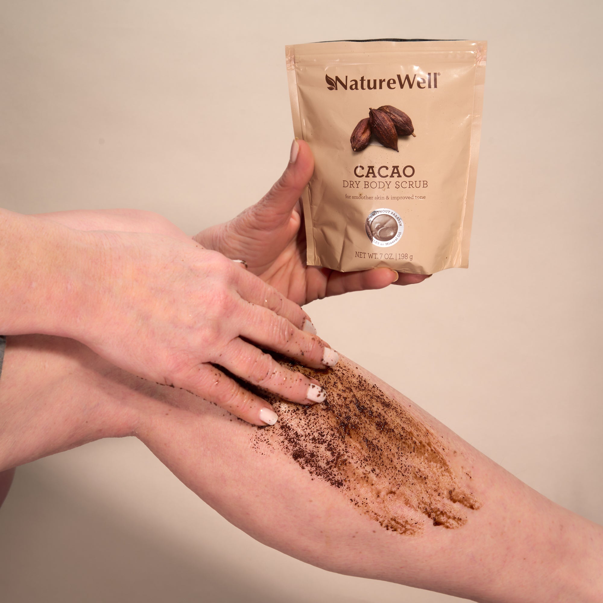 a woman's manicured hand applying brown colored cacao dry body scrub to her leg and holding the open bag of product in other hand