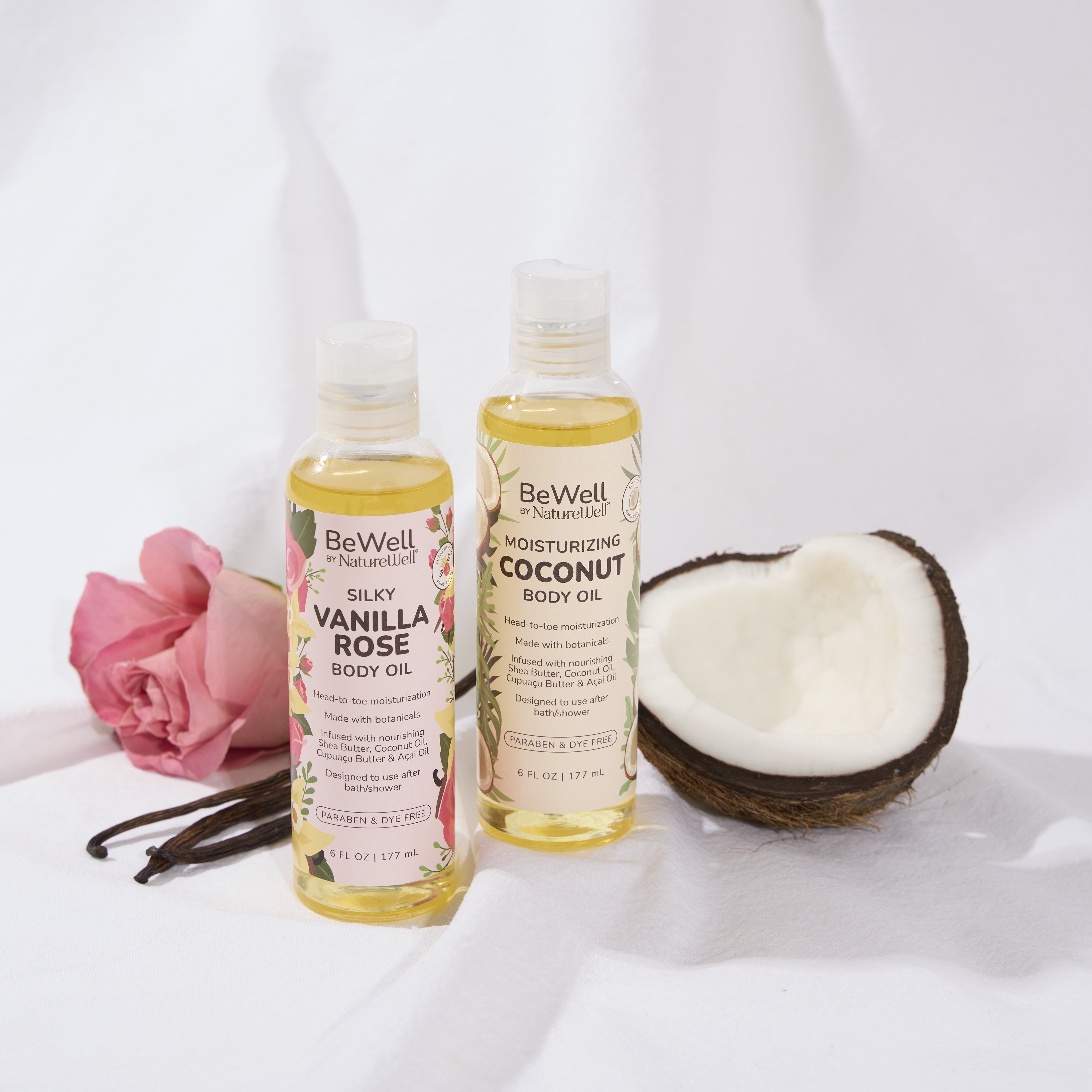 bottle of silky vanilla rose body oil and moisturizing coconut body oil next to an open coconut husk, pink rose and vanilla beans