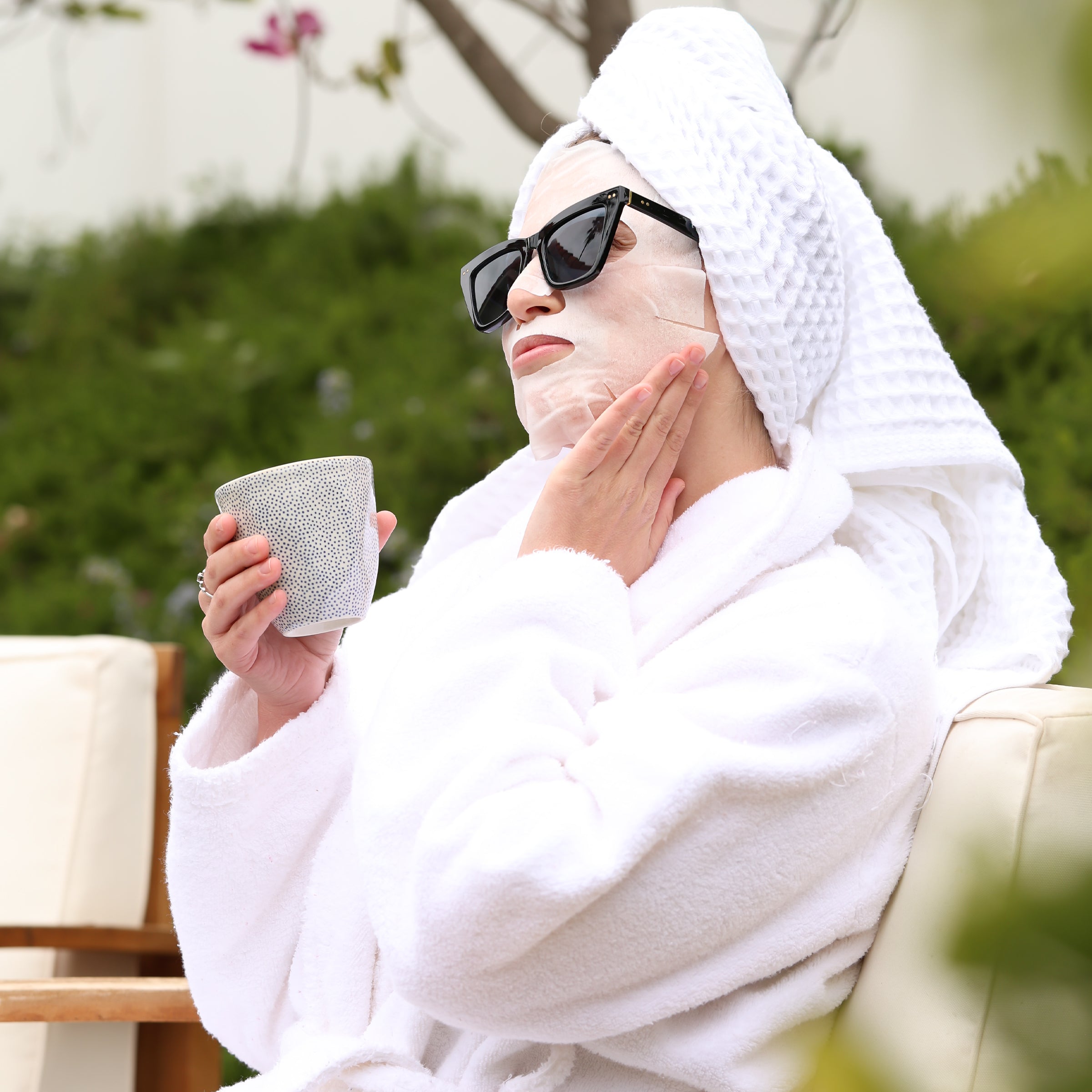 a woman relaxing in a spa setting wearing a hydrating sheet mask and sunglasses, dressed in a robe and holding a coffee mug